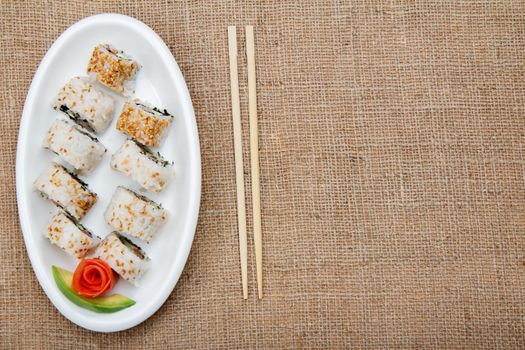 Uramaki sushi rolls with red fish and pickled ginger, piece of avocado on ceramic plate and wooden sticks on sackcloth. Top view with copy space