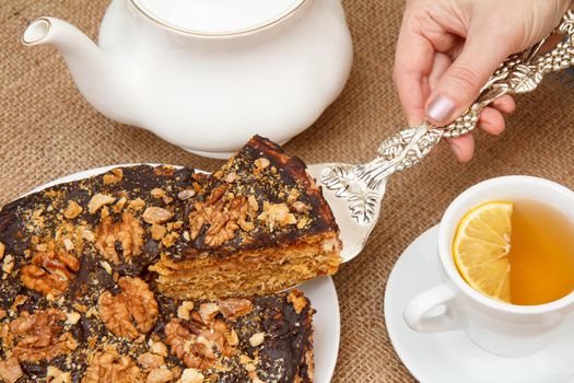 Cup of tea with slice of lemon, porcelain teapot and female hand holding piece of homemade chocolate cake with silver cake lifter