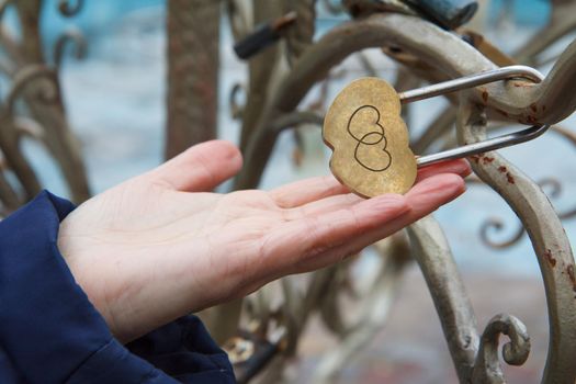 Female hand holds padlock with engraving of two hearts on metal tree. Selective focus on padlock