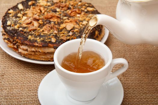 Tea is poured into a cup from porcelain teapot with homemade chocolate puff cake on the background. Selective focus on teapot