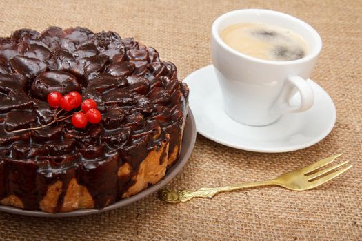 Chocolate cake decorated with bunch of viburnum, fork and cup of coffee beside it on table with sackcloth