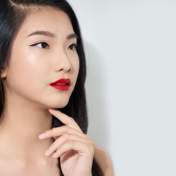 Beautiful stylish young woman is touching her chin, looking away and smiling, on white background