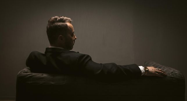 Thoughtful businessman sits at sofa in dark room. Rear view of pensive elegant man is alone with his thoughts. Lonelinessconcept. Leadership concept. Toned image.