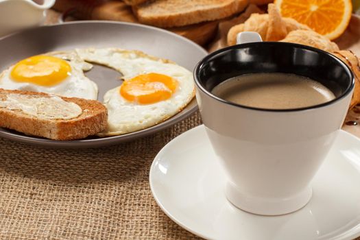 Cup of black coffee, plate with fried eggs and toast with butter, croissants, bread and fresh orange on table covered with sackcloth