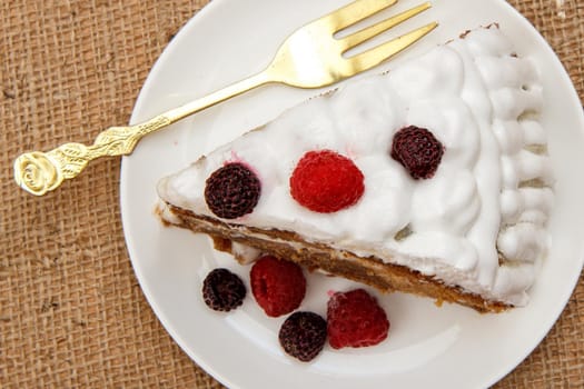 Slice of homemade biscuit cake decorated with whipped cream and raspberries and fork on table with sackcloth