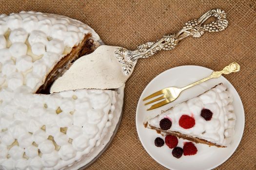 Sliced biscuit cake decorated with whipped cream and raspberries, silver cake lifter beside it on table with sackcloth. Top view