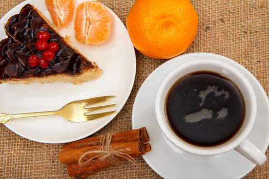 Slice of chocolate cake decorated with bunch of viburnum, fork, cup of coffee, oranges and cinnamon on table with sackcloth. Top view