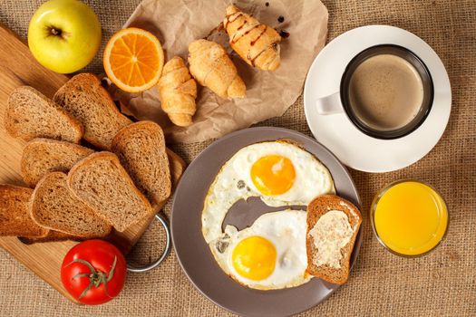 Top view of plate with fried eggs and toast with butter, glass of orange juice, cup of black coffee, croissants, bread on wooden cutting board, fresh tomato, apple on sackcloth