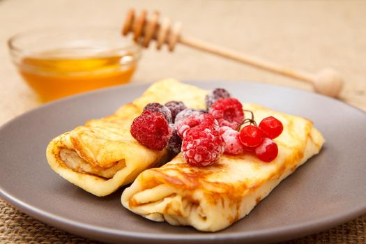 Homemade pancakes filled with cottage cheese and topped with frozen raspberries and blackberries on plate and honey in glass bowl with wooden spoon on background