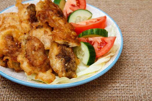 Plate with pieces of fried fish and freshly sliced tomatoes and cucumbers on table with sackcloth