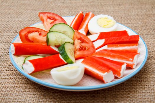 Plate with crab sticks, boiled egg and freshly sliced tomato and cucumber on sackcloth