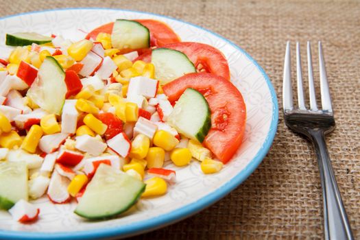 Salad with crab sticks, boiled corn and freshly sliced tomato and cucumber on plate with fork on sackcloth