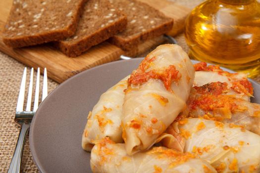 Delicious homemade cabbage rolls stuffed with rice and meat in plate, fork, bottle with sunflower oil and bread on wooden cutting board on sackcloth