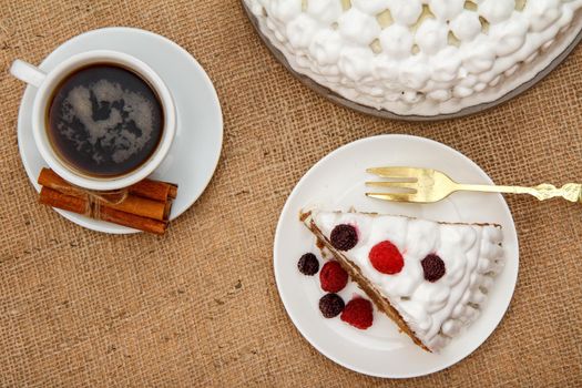 Cup of coffee, cinnamon, fork and slice of biscuit cake decorated with whipped cream and raspberries on table with sackcloth. Top view