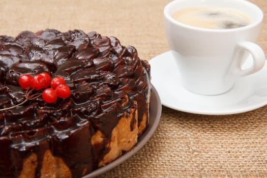 Chocolate cake decorated with bunch of viburnum and cup of coffee beside it on table with sackcloth