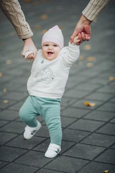 Portrait of cute sweet baby during walking outside. Parents holding their satisfied child by hands while it walking. Happy smiling infant in white sweater, green pants and white sneakers doing steps.