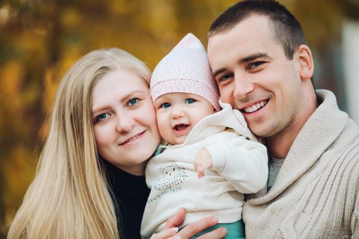 Portrait of attractive husband and blonde wife holding their surprised child. Handsome man helping pretty woman caring about little baby. Young happy family smiling at camera and standing together.