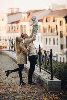 Side view of happy husband and wife with their lovely child. Handsome man holding little baby and beautiful woman hugging him from back. Young family in casual clothes walking together in sunny day.