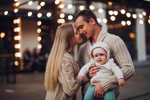 Portrait of young happy family standing together in cosiness atmosphere. Handsome man in sweater holding little smiling baby on hands. Blonde woman softly touching her hasband's face with closed eyes.