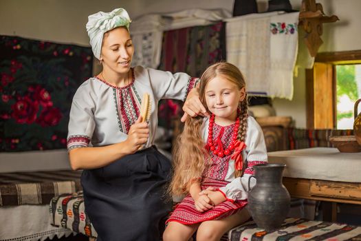 mother combing her daughter's hair, both dressed in Ukrainian national costumes