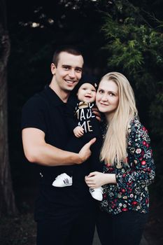 Portrait of lovely young family sitting together outside. Attractive blonde mother smiling with closed eyes. Handsome husband kissing his wife in head and holding happy baby in bright clothes.
