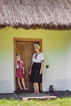mother and daughter in Ukrainian national costumes stand near the door from the house