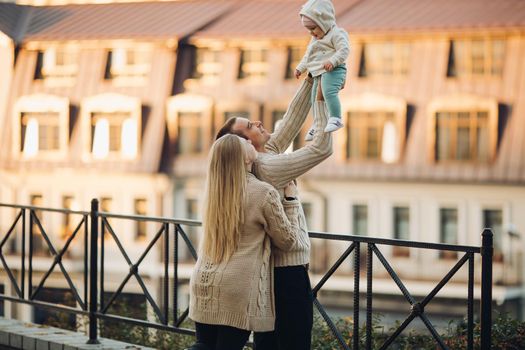 Side view of happy husband and wife with their lovely child. Handsome man holding little baby and beautiful woman hugging him from back. Young family in casual clothes walking together in sunny day.