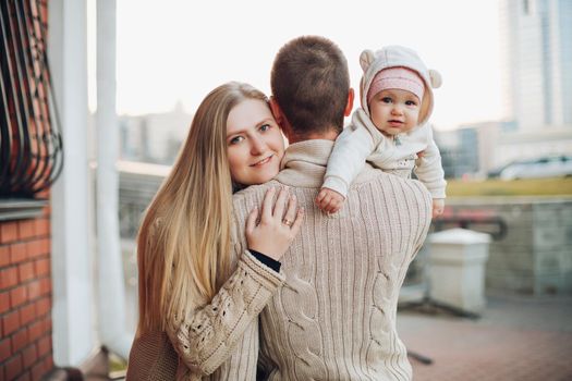 Portrait of attractive husband and blonde wife holding their surprised child. Handsome man helping pretty woman caring about little baby. Young happy family smiling at camera and standing together.