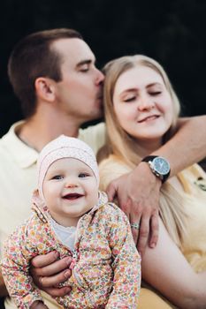 Portrait of lovely young family sitting together outside. Attractive blonde mother smiling with closed eyes. Handsome husband kissing his wife in head and holding happy baby in bright clothes.