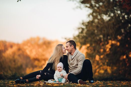 Portrait of lovely young family sitting together outside. Attractive blonde mother smiling with closed eyes. Handsome husband kissing his wife in head and holding happy baby in bright clothes.