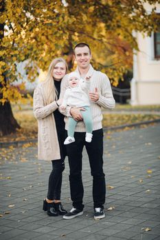 Portrait of lovely young family sitting together outside. Attractive blonde mother smiling with closed eyes. Handsome husband kissing his wife in head and holding happy baby in bright clothes.