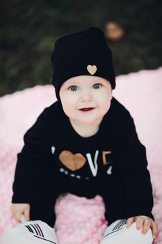 Little kid with cute big eyes lying on pink blanket on ground. Sweet and lovely child with big eyes looking at camera. Stylish infant in total black clothes and hat with gold heart leaning on hands.