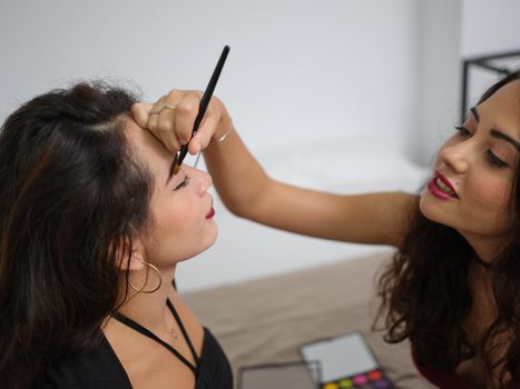 Cropped view of a young woman doing makeup eyebrow pencil to her female friend at home on halloween.