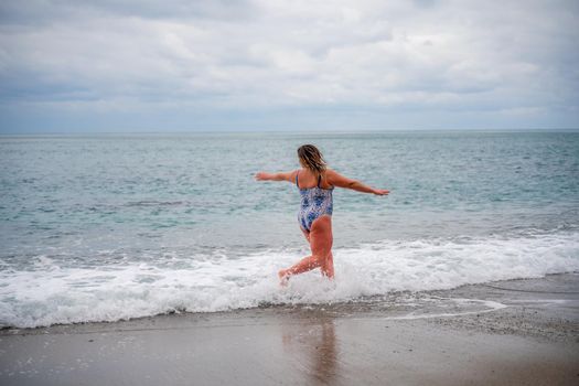 A plump woman in a bathing suit enters the water during the surf. Alone on the beach, Gray sky in the clouds, swimming in winter