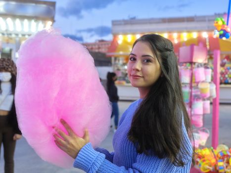 Woman turning to smile to the camera while holding a big pink cotton candy in a fair at night