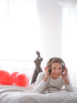 Vertical photo of smiling caucasian woman lying on the bed while listening to music with mobile