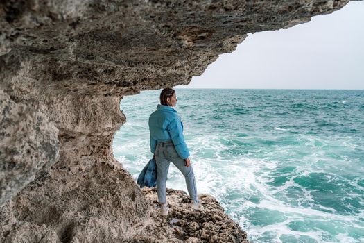 A woman in a blue jacket stands on a rock above a cliff above the sea and looks at the raging ocean. Girl traveler rests, thinks, dreams, enjoys nature. Peace and calm landscape, windy weather