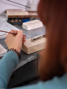 Vertical image of a woman working in her artisan jewelry workshop using a small brush on a ring to perform her work on a block of stone