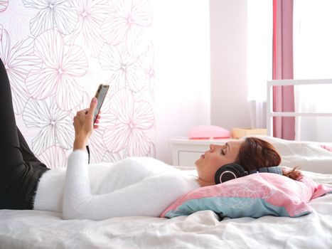 Profile of a woman watching a video on the mobile screen with headphones lying on the bed