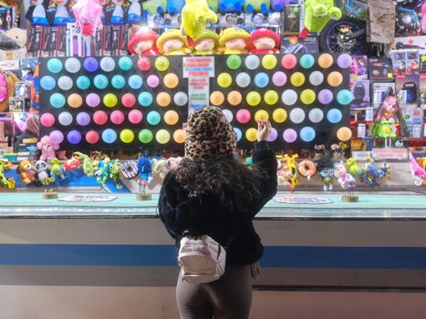 Back of a woman standing while throwing a dart in a colorful stall in a night-time fair