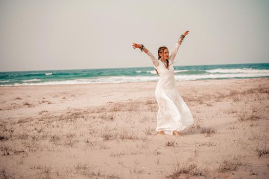 Model in boho style in a white long dress and silver jewelry on the beach. Her hair is braided, and there are many bracelets on her arms