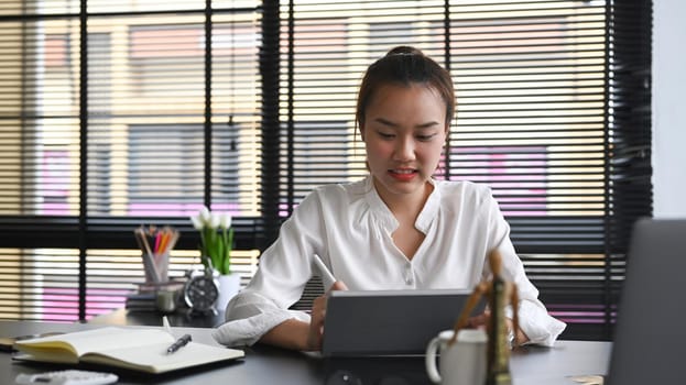Asian businesswoman sitting at modern workplace and using digital tablet.