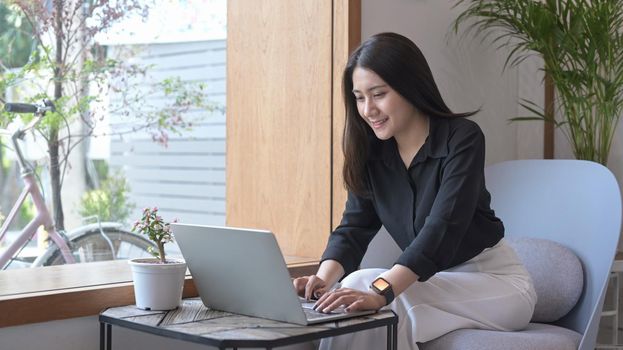 Pretty young female freelancer sitting on armchair and using laptop computer, working online, checking email.