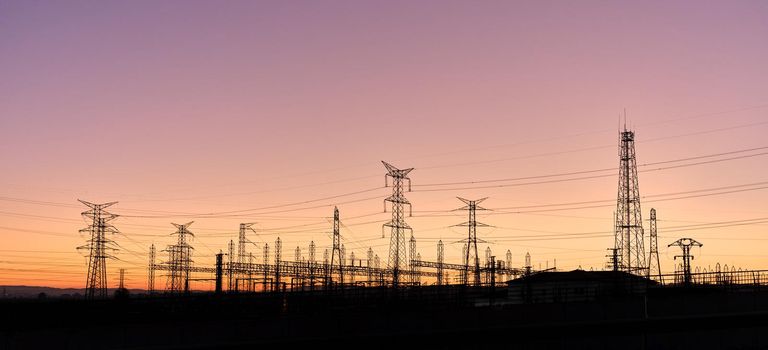 Panoramic photo of silhouettes of several electric towers connected by electric wires during sunset