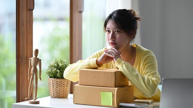 Thoughtful business woman selling online with cardboard boxes sitting in her home office.