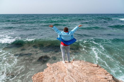 A woman in a blue jacket stands on a rock above a cliff above the sea and looks at the raging ocean. Girl traveler rests, thinks, dreams, enjoys nature. Peace and calm landscape, windy weather