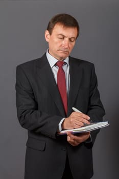 Confident and friendly elegant handsome mature businessman standing in front of a gray background holding a marker and writing in a notebook