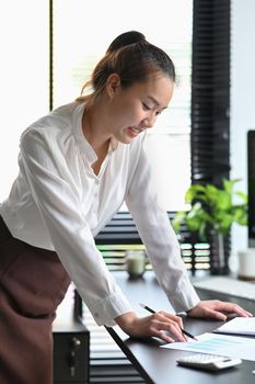 Asian female employee standing at her office desk and checking financial reports.