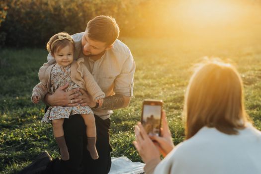 Young Family Having Picnic Outdoors, Woman Taking Pictures of Her Husband and Baby Daughter at Sunset