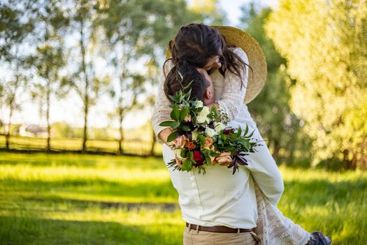 Stylish happy bride, groom hold picked up in hands and dancing. The groom raised and spins bride. Newlyweds kissing. On background on nature in the courtyard of house. Close up.